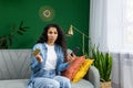 Upset cheated woman looking at camera, holding phone and bank credit card, sitting on sofa at home in living room