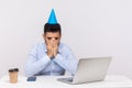 Upset depressed employee guy with party cone on head crying at workplace, hiding face feeling desperate lonely