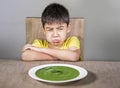 Upset and disgusted latin kid sitting on table refusing to eat spinach thick soup looking unhappy rejecting vegetarian food in