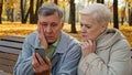Upset elderly married couple sit on bench in autumn park read bad news on smartphone worried old people look at screen Royalty Free Stock Photo