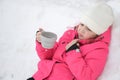 Upset frozen little cute girl in wool hat and pink ski suit drinking hot tea lying on white snow at cold winter holiday resort