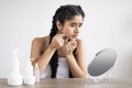 Upset Indian Woman With Problem Skin Looking At Pimple On Cheen While Sitting In Front Of Mirror At Home Royalty Free Stock Photo