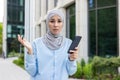 Upset islamic woman spreading hands in confusion while holding smartphone on urban background. Stressed lady in hijab