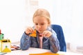 Upset little left-handed girl cutting orange color card at table