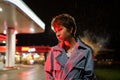 Upset lonely young millennial woman standing near petrol station shop in red neon light at night