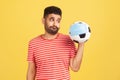 Upset bearded man in red t-shirt looking at soccer ball in safety face mask with disappointment, nervous because of football