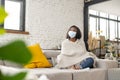 Upset multiracial woman with confirmed viral disease wearing medical mask feeling sad, sitting on the couch at home