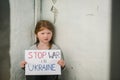 Upset poor toddler girl kid protesting war conflict raises banner with inscription text Stop war in Ukraine. Child sits near old