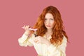 Upset pretty redhead girl posing with scissors and hair comb keeping in her hands and looking at the camera on pink Royalty Free Stock Photo