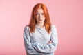Upset redhead girl with crossed arms in the studio on a pink background.