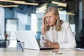 Upset senior businesswoman sitting at desk in office, holding head with hand and looking at laptop screen. Sad reads bad Royalty Free Stock Photo