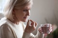 Upset senior woman holding pill and glass water taking medicine