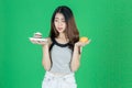 Upset stressed Asian woman choosing between orange fruit or unhealthy cake on hands over green isolated background. Healthy Royalty Free Stock Photo