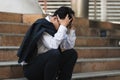 Upset stressed young Asian business man in suit with hands on head sitting on stairs. Unemployment and layoff concept