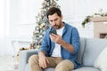 Upset unhappy man sitting near Christmas tree on winter day, got bad news on phone, celebrating new year and Christmas Royalty Free Stock Photo