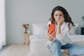 Upset puzzled female reading bad media news in social networks holding smartphone sitting in bedroom Royalty Free Stock Photo
