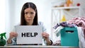 Upset woman showing help word on cardboard, laundry basket with clothes on table