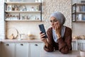 Upset and worried young muslim woman in hijab standing in the kitchen, leaning her hand on the table and looking at the Royalty Free Stock Photo