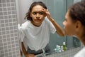 Upset young african woman finding pimple on her face while looking at mirror in bathroom home Royalty Free Stock Photo