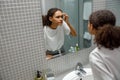 Upset young african woman finding pimple on her face while looking at mirror in bathroom home Royalty Free Stock Photo