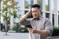 Upset young asian man standing near office building and holding head, reading bad news and message on phone Royalty Free Stock Photo