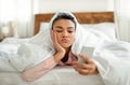 Upset young black woman looking at smartphone screen while laying in bed under blanket, feeling lonely or depressed Royalty Free Stock Photo