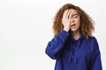 Upset young curly woman, student slap her face, facepalm and sighing disappointed, standing in blue hoodie over white