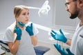 Upset young doctor`s assistant stand in dentistry room with dentist. She complain and point on cheeks. Guy look at woman