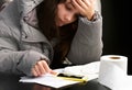 Upset young frozen woman puts on a hood sits at the table in cold room during turning off home heating in winter season