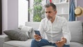 Upset young hispanic man, sporting grey hair and a beard, engrossed in a serious smartphone ordeal while comfortably nestled on