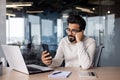 Upset young Indian male businessman sitting at desk in office leaning tiredly on hand and looking frustrated at phone Royalty Free Stock Photo