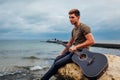Upset young man holding acoustic guitar on beach surrounded with rocks on rainy day. Feeling lonely and sad Royalty Free Stock Photo