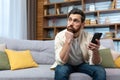 An upset young man is sitting on the sofa at home with a phone in his hands, looking thoughtfully to the side. Waiting Royalty Free Stock Photo