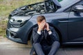 Upset young man use phone sitting on road near the broken car opened the hood help repair Royalty Free Stock Photo