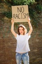 Upset young protesting woman in white shirt holds protest sign broadsheet placard with slogan `No racism` for public