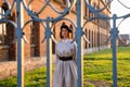 Upset young woman in black beret and grey dress stands near the old brick wall behind the old forged fence Royalty Free Stock Photo