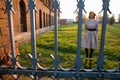 Upset young woman in black beret and grey dress stands near the old brick wall behind the old forged fence Royalty Free Stock Photo