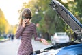 Upset young woman driver talking angrily on cell phone with assistance service near a broken car with open hood on a city street Royalty Free Stock Photo