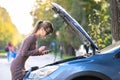 Upset young woman driver talking angrily on cell phone with assistance service near a broken car with open hood on a city street Royalty Free Stock Photo