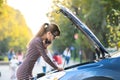 Upset young woman driver talking angrily on cell phone with assistance service near a broken car with open hood on a city street Royalty Free Stock Photo