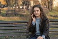 Upset young woman with long wavy brown hair sits on bench in park talks on phone. Woman in autumn park Royalty Free Stock Photo