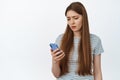 Upset young woman looking at her mobile phone, reading smartphone message disappointed, standing over white background Royalty Free Stock Photo