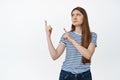 Upset young woman pointing and looking at upper left corner with skeptical, disappointed face expression, standing over