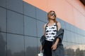 Urban attractive young hipster woman in stylish clothes in vintage sunglasses with a backpack poses in a city near a modern gray Royalty Free Stock Photo