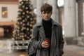 Urban attractive young man is standing in a stylish plaid coat with a trendy hairstyle with a backpack with hot coffee in hands