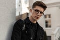 Urban fresh portrait confident young man with hairstyle in fashion black denim jacket in stylish glasses near wall outdoors.