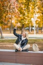 Urban golden autumn. Adult woman sits a parapet in park and chill out playing with a bouquet of fallen leaves, outdoor