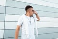 Urban handsome young hipster man in a trendy white t-shirt in stylish sunglasses with fashionable hairstyle posing near a modern Royalty Free Stock Photo