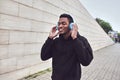 urban happy smiling african man in wireless headphones enjoying listening to music wearing black hoodie on city street