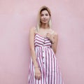 Urban happy stylish young woman blonde with cute smile in vintage striped long dress posing near a pink building on the street